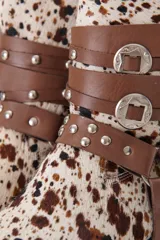 Bota de cuero color marrón con puntera fina y taco bajo. Presenta correas de cuero negro con apliques metálicos plateados.