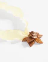 Collar con cinta de tela plisada color amarillo pálido, atado al cuello, con un dije colgante que simula una flor de hibisco en tonos marrones y dorados, con un detalle central perlado.