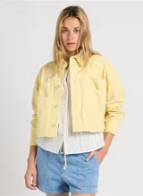 Chaqueta casual color amarillo pastel, de corte corto y suelto. Tiene cuello camisero, cierre frontal con botones metálicos, mangas largas y grandes bolsillos de parche en la parte delantera.