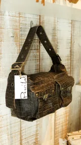 Bolso de hombro color beige con textura de cuero grabado tipo cocodrilo. Presenta dos bolsillos frontales con solapa y cierres metálicos, y una correa ancha ajustable con ojales metálicos.