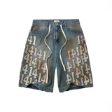 Shorts de jean en color azul con efecto desgastado, manchas de pintura y apliques bordados con números y letras góticas. Presenta un corte holgado (baggy) y cordones decorativos en la cintura.