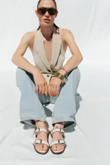 Sandalia plana de color beige con dos tiras ajustables mediante hebillas metálicas doradas. Presenta un diseño de punta redonda y suela plana con borde en contraste.