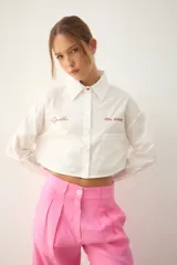 Camisa blanca de popelina de algodón, de corte cropped, con cuello clásico y cierre frontal de botones. Presenta bordados rojos en el frente con las frases "el poder" y "DEL AMOR", y un bolsillo de parche en el lado derecho.