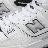 Championes de caña alta New Balance modelo 650, con diseño en cuero blanco, detalles en negro y refuerzos de gamuza gris en la puntera. Presenta el logo de la marca en el lateral y suela de goma con tecnología de amortiguación.