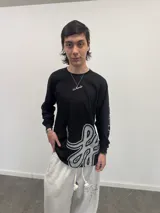 Remera manga larga de algodón color negro, con diseño gráfico abstracto en tono gris en la parte inferior frontal y tipografía cursiva en la manga. Presenta cuello redondo reforzado y grifa de la marca en el puño.