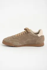 Zapatilla deportiva de gamuza color visón, con cordones a tono y suela de goma marrón. Presenta un diseño retro de running con el nombre de la marca grabado en dorado en el lateral.