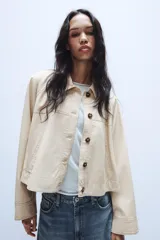 Trench corta de color beige, con cuello solapa, manga larga y cierre frontal con botones oscuros. Presenta bolsillos de vivo en el delantero y un diseño de corte recto.