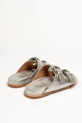 Sandalias de cuero color negro con dos tiras anchas sobre el empeine, cada una con hebillas metálicas ajustables y detalles de tachas plateadas. La suela es de cuero con un borde de color madera.