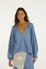 Blusa de mezclilla ligera con escote en V pronunciado, mangas largas amplias con puños ajustables mediante cordón y detalles de tiras colgantes en el cuello.