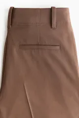 Pantalón de corte holgado color marrón, de estilo sastrero, con cintura clásica, trabillas para cinto y cierre con botón y cremallera. Presenta piernas rectas con pinzas frontales y bolsillos laterales.