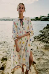 Vestido largo estilo túnica de color crudo con estampado multicolor de motivos marinos y mediterráneos. Presenta cuello con volante, cierre frontal con botones, manga larga y cordón ajustable en el cuello con abalorios.