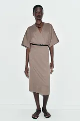 Vestido midi de tejido piqué en color marrón oscuro jaspeado, con escote en pico cruzado y mangas cortas. Presenta detalle de frunces en la cintura y un cinturón integrado con hebilla metálica a contraste.