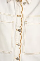 Chaleco de denim blanco con diseño sin mangas, cuello camisero, bolsillos frontales con solapa y cierre de botones metálicos con detalle de ribete ondulado en contraste.