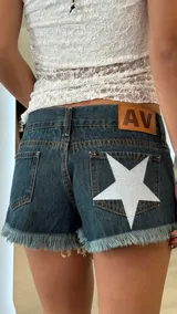 Short de jean azul oscuro con ruedo deshilachado, tiro bajo y detalle de estrella grande estampada en color blanco brillante sobre el bolsillo trasero derecho. Incluye etiqueta de cuero con logo AV en la parte posterior.