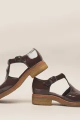 Zapato tipo guillermina de cuero charol color marrón oscuro, con tira T y hebilla ajustable en el lateral. Posee puntera cuadrada y suela de goma color caramelo con taco bajo.