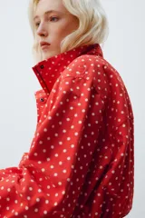 Chaqueta roja con estampado de lunares blancos, cuello subido, manga larga con puños plisados, bolsillos delanteros, bajo ajustable con cordón y cierre frontal con cremallera.