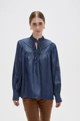 Blusa de lyocell en tono azul denim, con diseño holgado y frunces en el canesú. Presenta cuello alto con volados, cierre frontal mediante cordón ajustable y mangas largas con puños abotonados.
