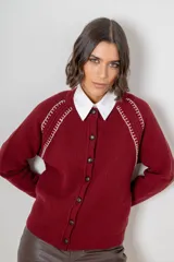 Saco de punto color beige, con cierre frontal de botones marrones y detalle de costuras decorativas oscuras en los hombros. Presenta un diseño clásico con cuello redondo.