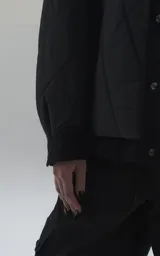 Campera tipo sobrecamisa de diseño oversize, confeccionada en microfibra opaca con patrón matelaseado geométrico. Presenta cuello, puños, cartera central y ruedo en gabardina de algodón, con cierre frontal mediante broches a presión. El diseño del ruedo simula una prenda superpuesta.