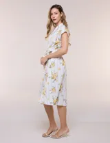 Vestido camisero midi de silueta evasé, confeccionado en tejido liviano blanco con estampado floral amarillo y rayas celestes. Cuenta con cuello solapa en V, cartera con botones frontales, mangas cortas japonesas y cintura definida con lazo ajustable a juego.