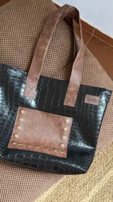 Bolso tote negro con textura símil cuero de cocodrilo y bolsillo frontal marrón con tachas plateadas. Tiene doble asa de hombro marrón y cierre superior.