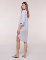 Vestido camisero corto con estampado a rayas verticales en color marrón y blanco, de cuello camisero con escote en V y botones frontales parciales. Cuenta con bolsillos laterales y en el pecho, mangas largas con presillas para ajustar y ruedo curvo asimétrico.