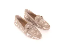 Mocasín de gamuza color avena con bordados florales en hilo blanco y pequeños apliques metálicos dorados. Presenta un lazo decorativo en la capellada y suela de goma flexible.