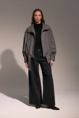 Campera tipo bomber de corte holgado y diseño estructurado, confeccionada en tejido gris con textura de espiga. Presenta cuello alto con cierre de broches a presión, mangas largas con puños ajustados y un cinturón integrado en el ruedo con hebilla metálica para un ajuste personalizado.