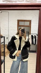 Campera tipo gamulan de gamuza negra con interior y bordes de corderito sintético color crema. Presenta detalles de tiras en cuero marrón, cierre frontal y hebillas ajustables en los laterales.