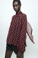 Camisa de cuello subido y manga larga con puños elásticos. Presenta un diseño de lunares en tonos fucsia y amarillo sobre fondo negro, con detalle de pliegues en el frente y cierre de cremallera oculta en la espalda.