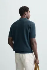 Polo de manga corta color verde azulado oscuro, confeccionado en tejido de punto con textura. Presenta cuello solapa y cierre frontal de botonadura.