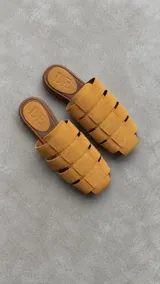 Zuecos de punta cuadrada confeccionados en gamuza color mostaza, con diseño de tiras cruzadas y suela plana.