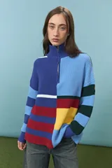 Sweater de tejido de punto grueso con diseño de bloques de color y rayas horizontales en tonos azul, rojo, amarillo y verde. Presenta cuello alto con cierre frontal parcial y mangas largas con puños ajustados.