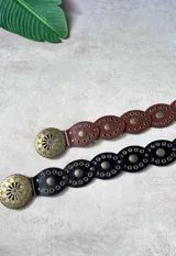 Cinto ancho de cuero sintético con diseño de eslabones ovalados, cada uno adornado con múltiples ojales metálicos y un remache central con diseño floral. Hebilla grande redonda con diseño calado de estilo tribal o western. Disponible en color negro y marrón rojizo.