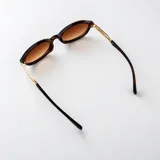 Lentes de sol con armazón ovalado estilo carey (tortoise) en tonos marrones y negros, con detalles metálicos dorados en las patillas y lentes degradados en tono marrón.