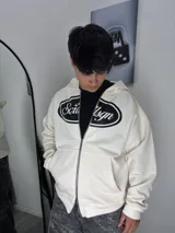 Campera tipo hoodie con cierre frontal completo, color beige, con estampado gráfico ovalado en el pecho y bolsillos tipo canguro.