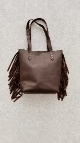 Cartera tipo tote de cuero marrón, con tiras de flecos colgantes en los laterales y doble asa superior para el hombro.