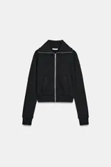 Chaqueta de felpa negra con cuello solapa, cierre frontal de cremallera metálica, mangas largas con puños elásticos y bolsillos de plastrón en el delantero.