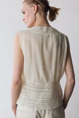 Blusa sin mangas de cuello redondo, confeccionada con detalles de encaje y puntilla a tono. Presenta cierre frontal con botones y diseño estructurado.