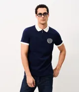 Polo de manga corta color azul marino, confeccionado en jersey de algodón texturizado. Cuenta con cuello polo en contraste de color blanco con cierre frontal parcial y ribetes blancos en los puños. Presenta un escudo bordado en el pecho.