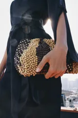 Bolso tipo pouch confeccionado completamente con cuentas de madera en tonos marrón, beige y tostado, formando un patrón abstracto. Cuenta con una correa fina de cadena dorada desmontable y cierre superior con cremallera.