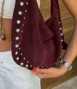 Bolso de hombro tipo hobo color negro, con acabado de gamuza o material similar. Decorado con tachas metálicas redondas plateadas a lo largo del borde inferior y en un dije colgante con forma de corazón.