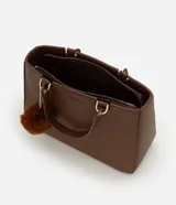 Cartera estilo satchel de tamaño mediano, confeccionada en poliuretano color marrón con textura tipo cuero. Cuenta con asas de mano reforzadas, correa larga desmontable, cierre superior de cremallera y un colgante decorativo de pompón de pelo sintético.