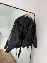 Campera estilo biker oversize de cuero sintético negro, con cierre frontal cruzado, solapas grandes, hombreras y cinturón ajustable en la cintura con hebilla metálica.