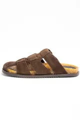Sandalias tipo zueco de cuero gamuzado color marrón, con diseño de tiras cruzadas, puntera cerrada y una gran hebilla metálica dorada en el lateral. Cuenta con base de corcho y suela plana.