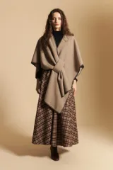 Poncho de tejido suave color beige con diseño envolvente y terminaciones con costuras decorativas en contraste color negro. Presenta un detalle de lazo frontal integrado.