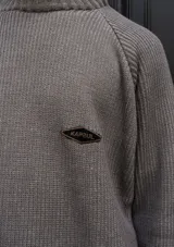 Buzo de tejido de punto con textura acanalada en color negro con detalles en verde oliva. Presenta cuello redondo, mangas largas tipo raglán y un pequeño parche decorativo en el pecho. El diseño incluye un borde inferior con franjas horizontales en tono contrastante.