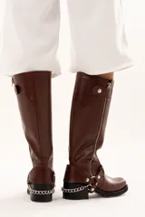 Bota de caña alta estilo biker confeccionada en cuero color chocolate. Presenta un cierre metálico lateral que recorre toda la caña, taco cuadrado y plataforma. Incluye un sistema de estribo y cadena metálica desmontable en la zona del tobillo, permitiendo diversas formas de uso.
