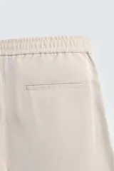 Pantalón de hombre de corte relajado, color beige claro, con cintura elástica, cierre frontal con botón y cremallera, bolsillos delanteros y bolsillos traseros de vivo.