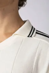 Remera tipo chomba de tejido de punto color blanco, con cuello polo y diseño de rayas verticales sutiles. Presenta estampado gráfico frontal con texto y logo, además de detalles de rayas negras en los hombros.
